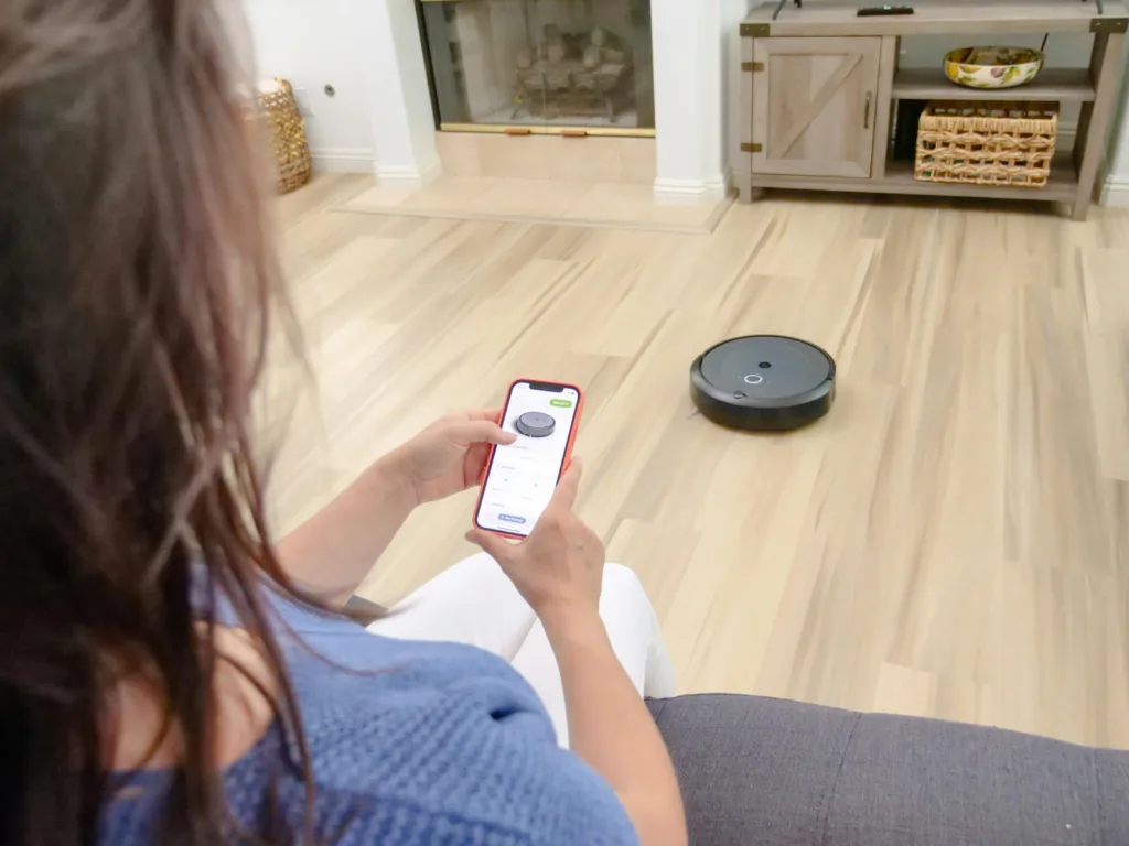 A woman uses her smartphone to control a Roborock Saros Z70 robot vacuum with arm navigating across light wood flooring, illustrating the ease of use of the app. The scene highlights the automation of household cleaning and remote control of this smart robot vacuum cleaner.