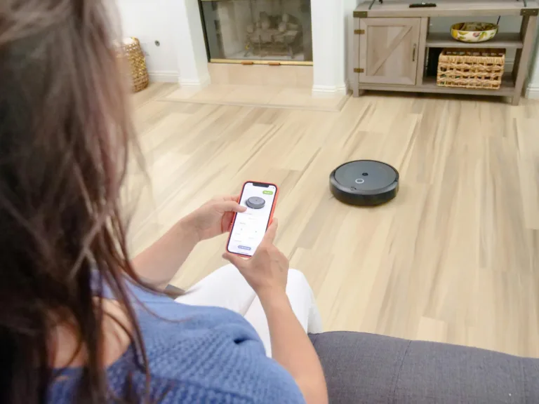 A woman uses her smartphone to control a Roborock Saros Z70 robot vacuum with arm navigating across light wood flooring, illustrating the ease of use of the app. The scene highlights the automation of household cleaning and remote control of this smart robot vacuum cleaner.
